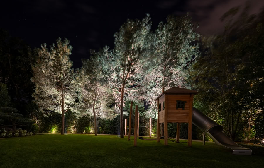 Aire de jeux en bois avec toboggan, entourée d'arbres éclairés la nuit à Versoix.