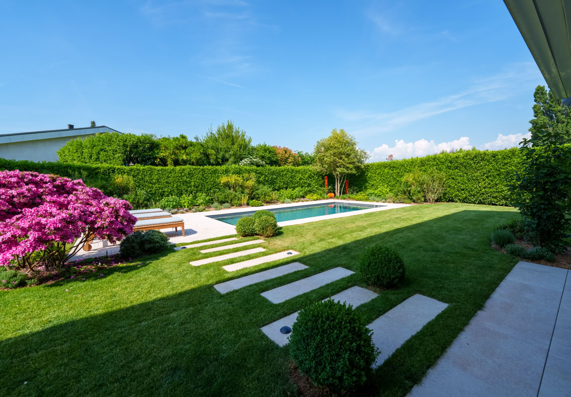 Réalisations. Jardin avec piscine, pelouse verte, arbustes taillés, fleurs roses à gauche, ciel bleu.