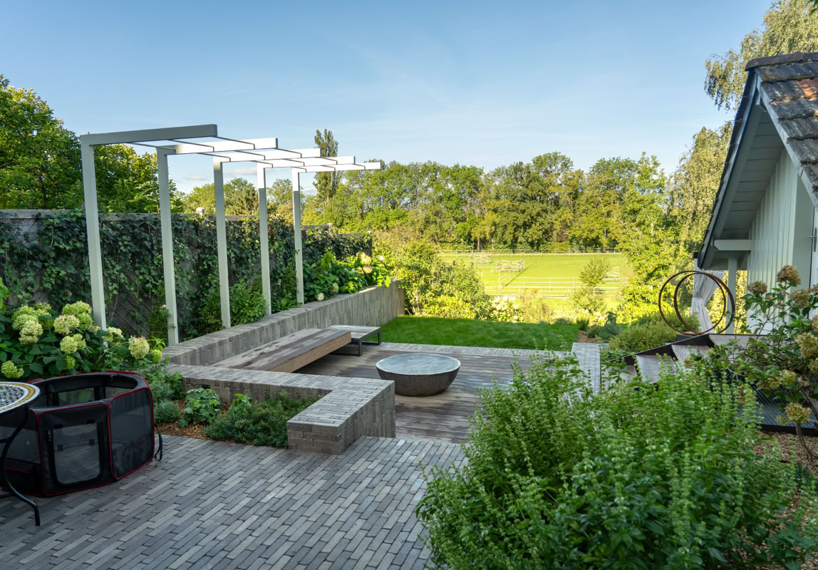 Terrasse avec des bancs en béton, un foyer, des plantes vertes, et vue sur un jardin à Meinier avec pelouse et arbres.