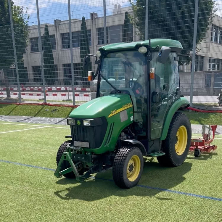 Tracteur vert sur un terrain de sport en gazon synthétique, avec des bâtiments en arrière-plan.