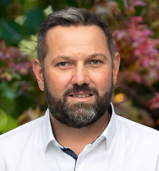 Arnaud Kohlmann souriant portant une chemise blanche, avec un fond flou de feuillage coloré.