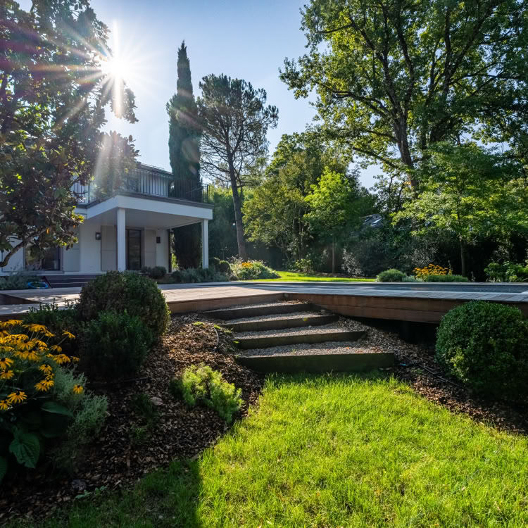 Maison blanche entourée de verdure avec un jardin bien entretenu, des fleurs jaunes, des buissons et des arbres, éclairée par le soleil.