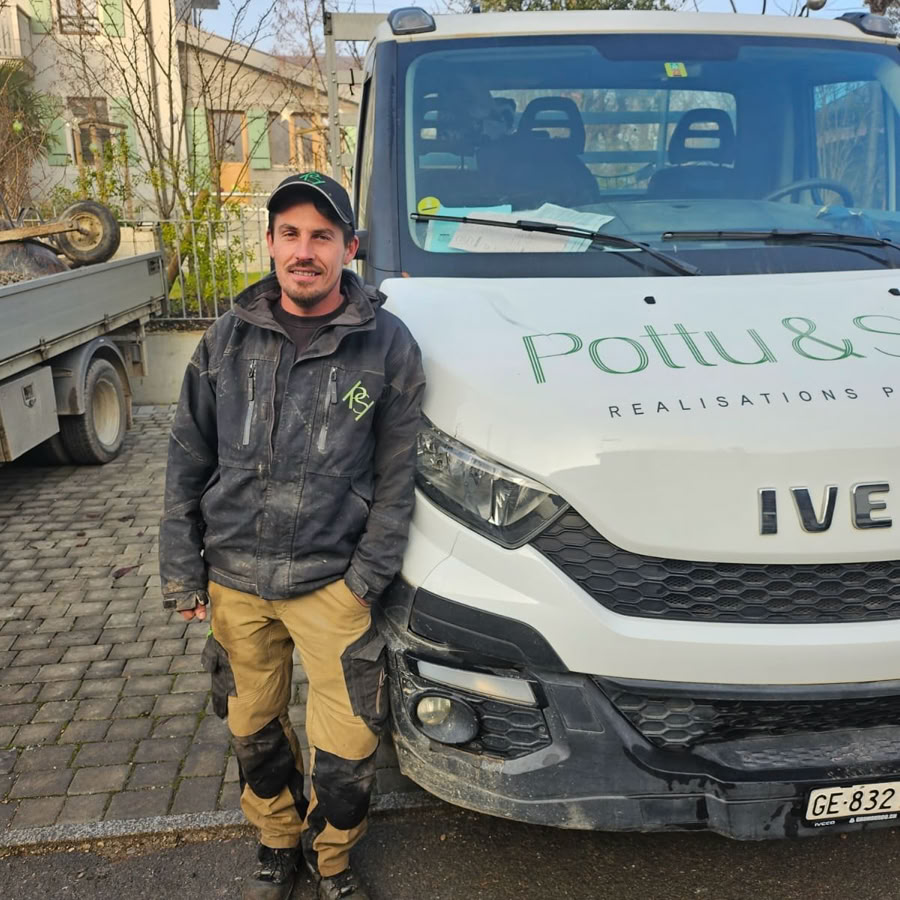 Carrière. Candido, Maçon paysagiste chez Pottu & Seitz en tenue de travail se tient à côté d'une camionnette sur une route pavée, avec des bâtiments et des arbres en arrière-plan.