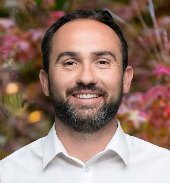 Romain Revillon souriant portant une chemise blanche, avec un fond flou de feuillage coloré.