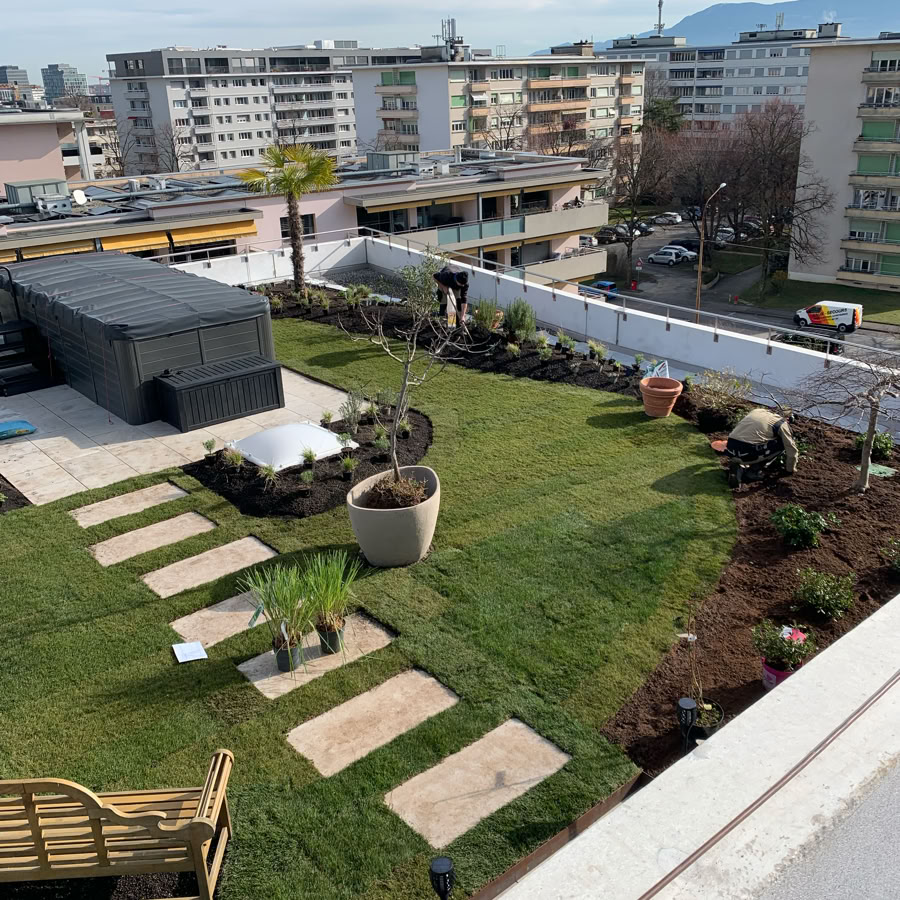 Jardin en toiture à l'aéroport de Genève avec des arbustes taillés et des plantes basses, bordé par un trottoir en béton, sous un ciel bleu.