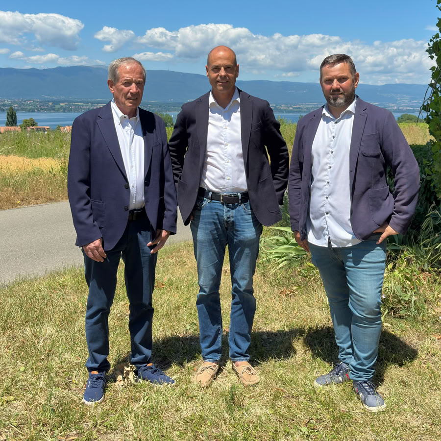 A propos de nous. Alain Richner, Jean-Jacques Seitz, Arnaud Kohlmann debout sur une pelouse près d'une route, avec un lac et des montagnes en arrière-plan sous un ciel bleu.
