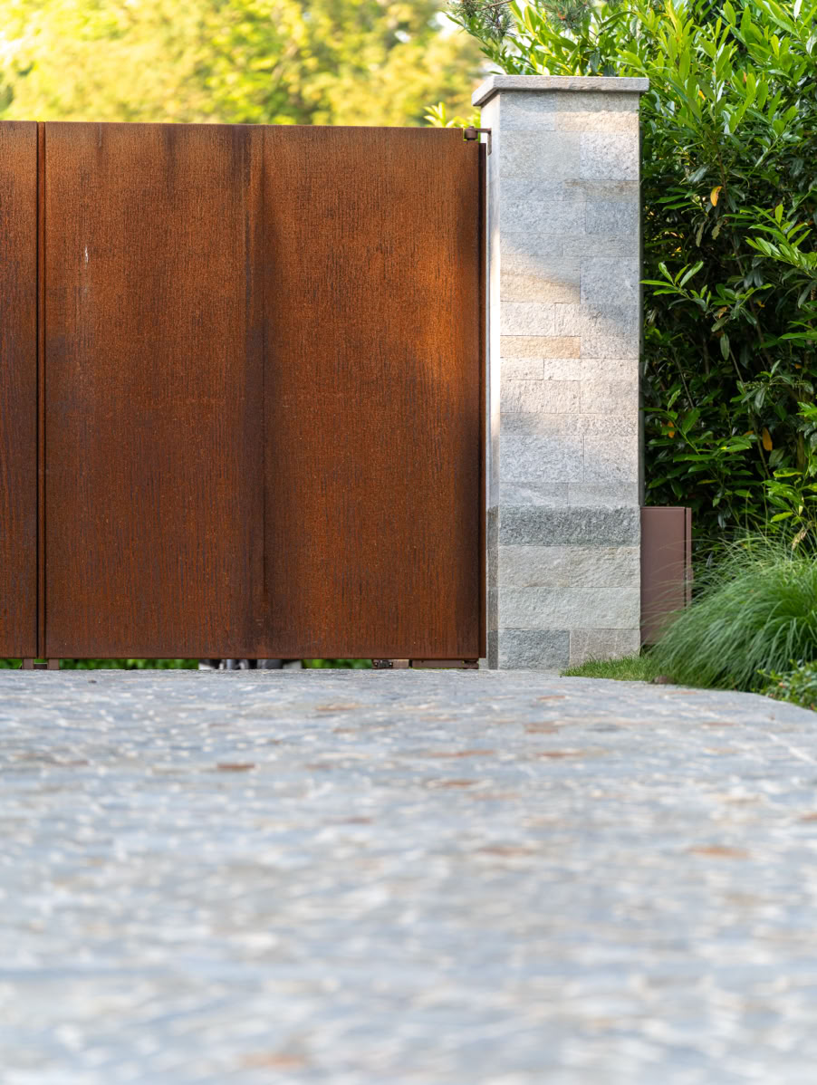 Allée pavée menant à un portail en métal rouillé avec un pilier en pierre, entouré de verdure.