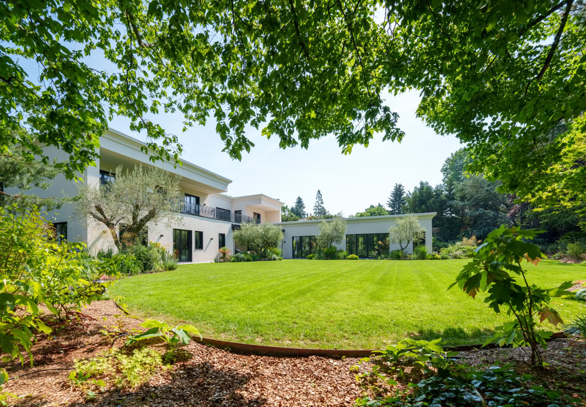 Grande maison blanche à Collonge-Bellerive avec un jardin verdoyant, entourée d'arbres et de buissons.