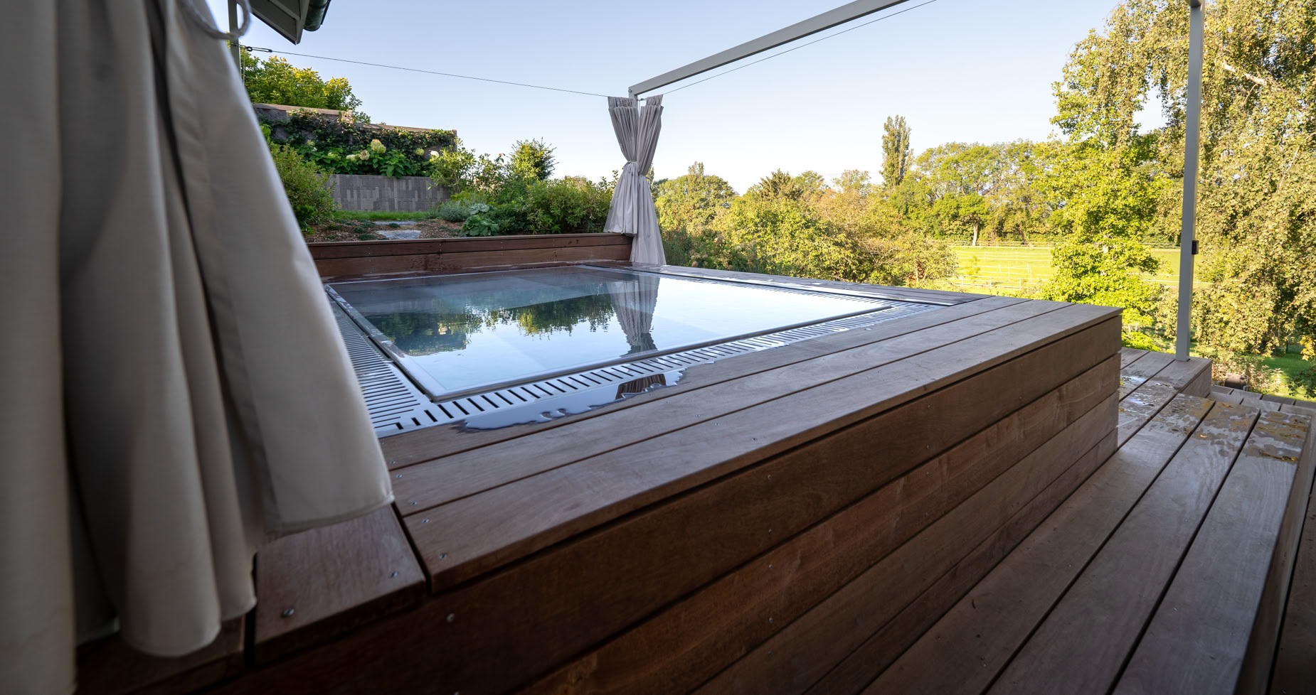 Terrasse en bois avec un jacuzzi encastré, entourée de rideaux et offrant une vue sur un aménagement de jardin à Meinier verdoyant.