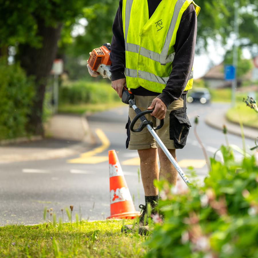 Jardinier Pottu & Seitz portant un gilet de sécurité jaune utilisant un taille-bordures près d'une route, avec un cône de signalisation orange et blanc à proximité.