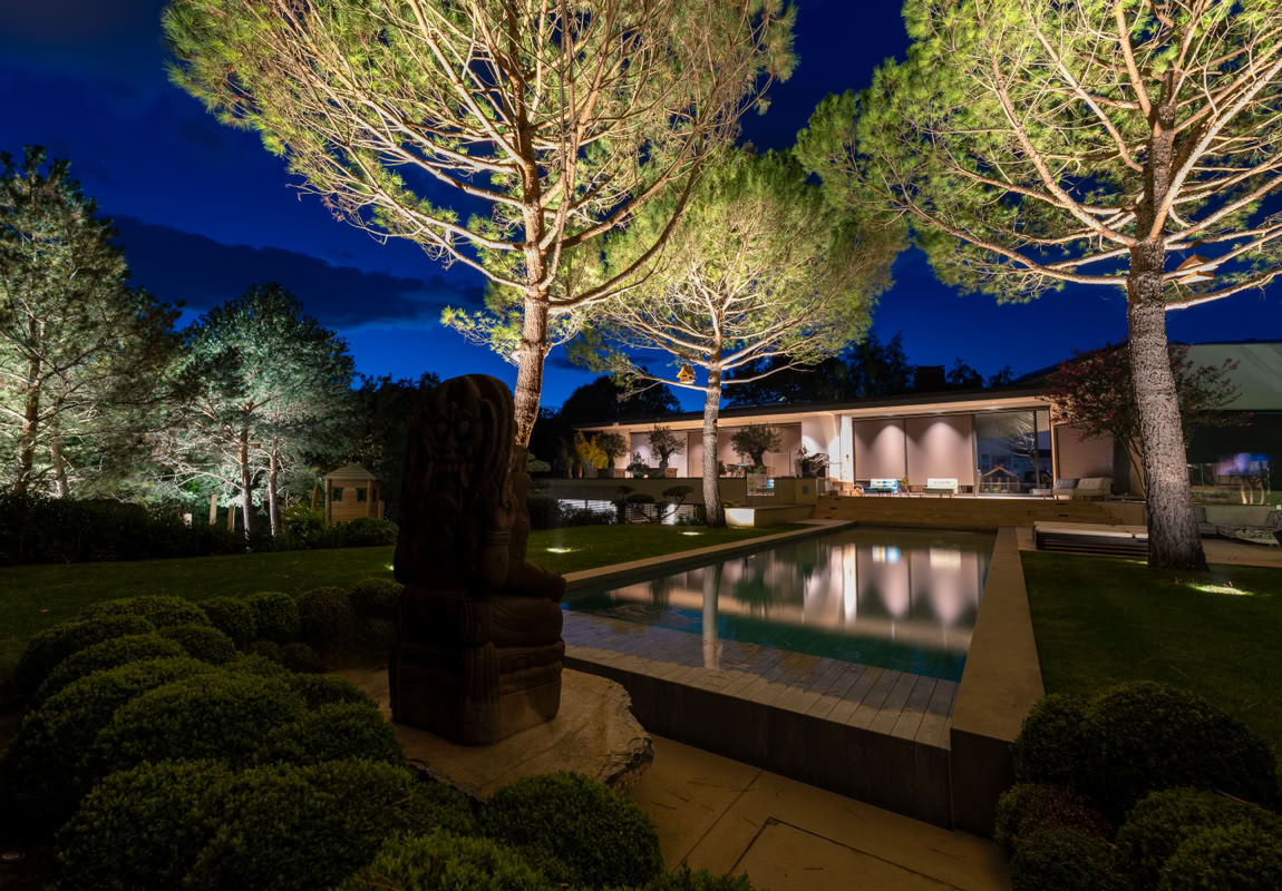 Jardin à Versoix éclairé la nuit avec des arbres, une piscine et une maison moderne en arrière-plan.