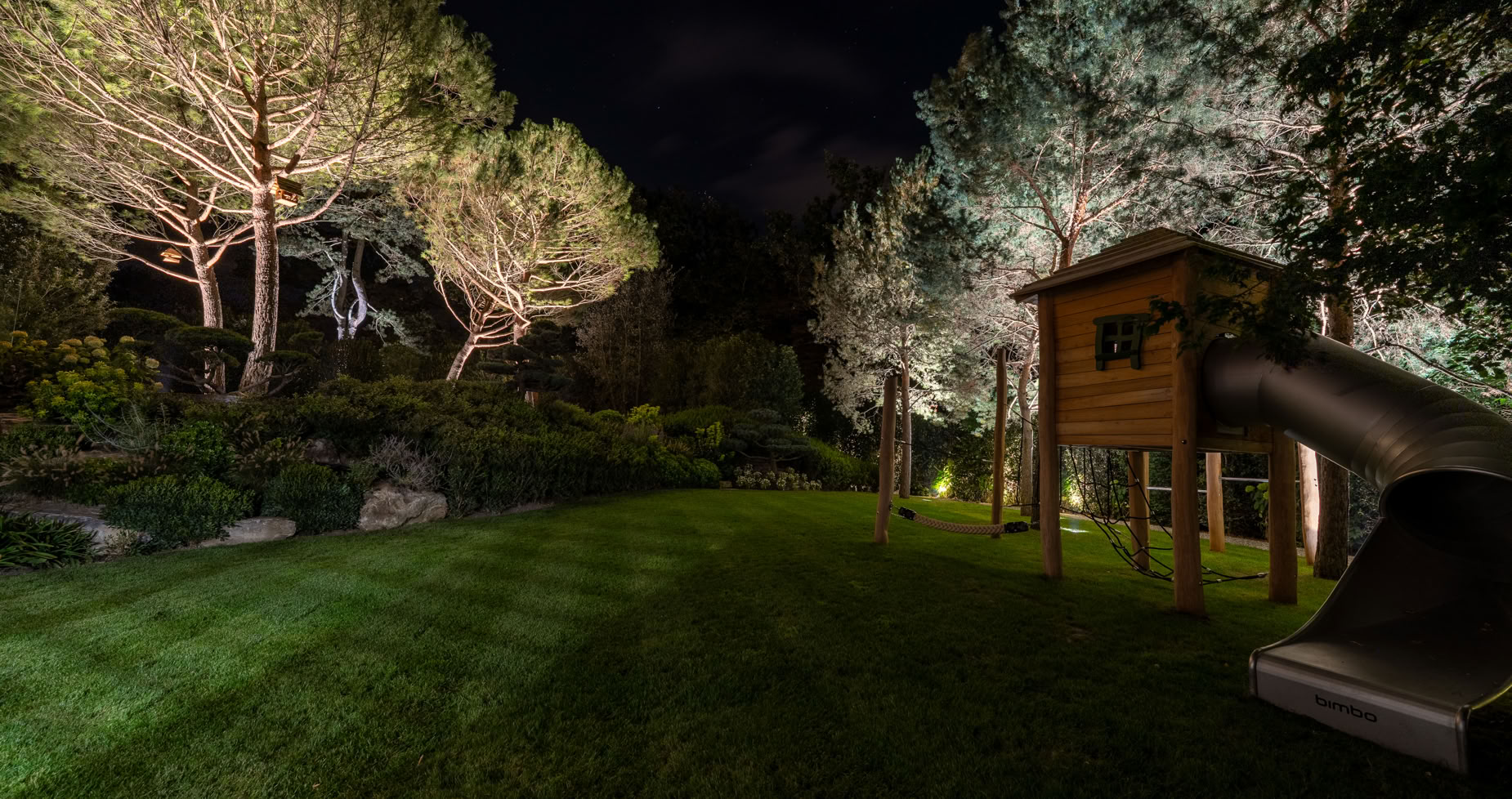 Pelouse éclairée la nuit avec des arbres en arrière-plan et une cabane en bois avec un toboggan à droite.