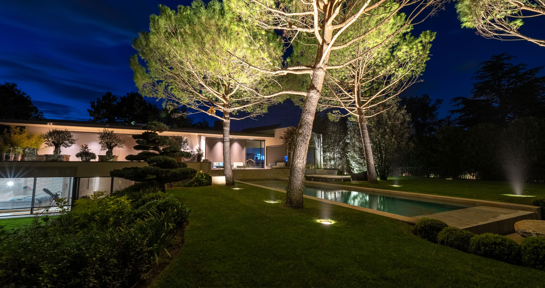 Vue nocturne d'un jardin à Versoix avec des arbres éclairés, une pelouse bien entretenue, une piscine et un bâtiment moderne en arrière-plan.
