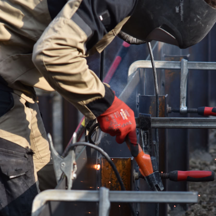 Paysagiste Pottu & Seitz portant un casque de protection et des gants rouges, utilisant un outil de soudage sur des structures métalliques.