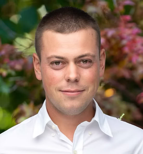 Florian Vidonne souriant portant une chemise blanche, avec un fond flou de feuillage coloré.