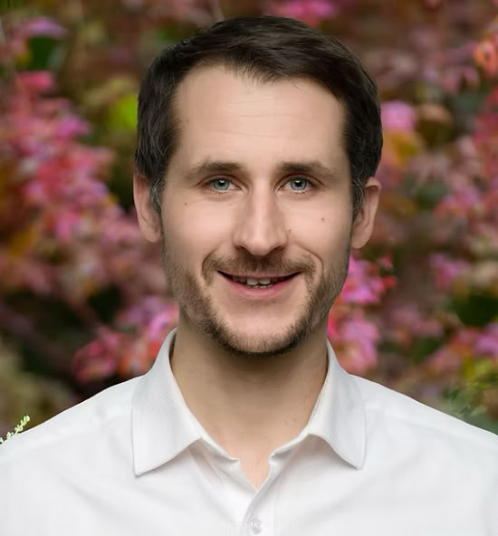 Jérémy Fricaud souriant portant une chemise blanche, avec un fond flou de feuillage coloré.