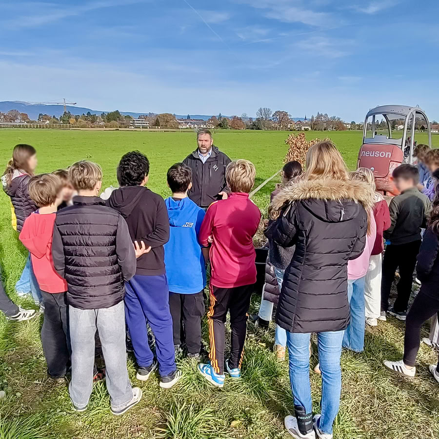 Un groupe d'élèves debout dans un champ vert, écoutant un employé de chez Pottu & Seitz qui parle devant eux, avec un ciel bleu en arrière-plan.
