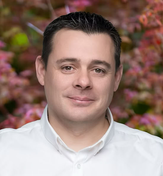 Lionel Lachavanne souriant portant une chemise blanche, avec un fond flou de feuillage coloré.