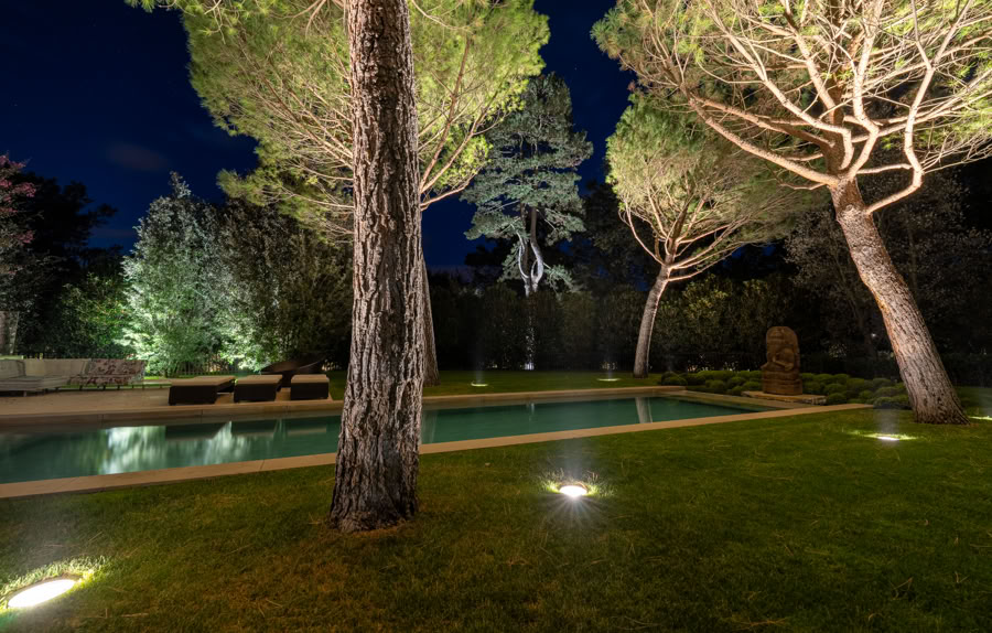 Piscine extérieure entourée d'arbres éclairés la nuit, avec pelouse et chaises longues.
