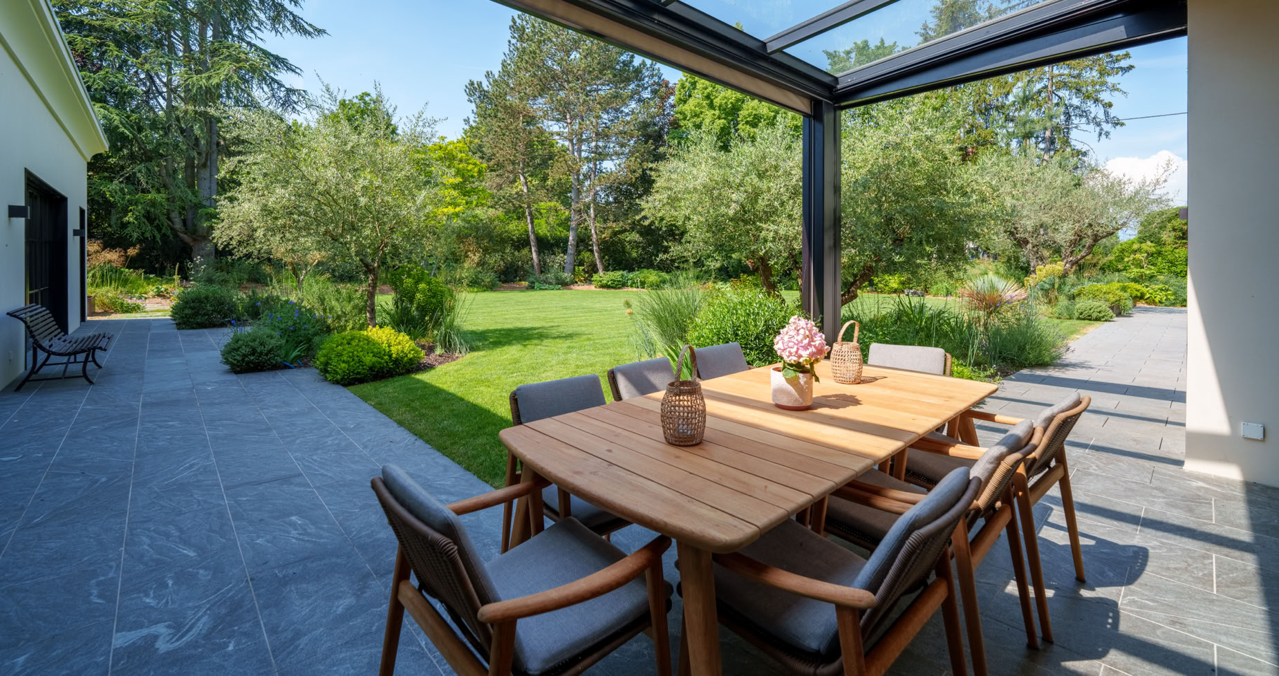 Terrasse avec une table en bois entourée de chaises, sous une pergola en verre, donnant sur un jardin verdoyant à Collonge-Bellerive.