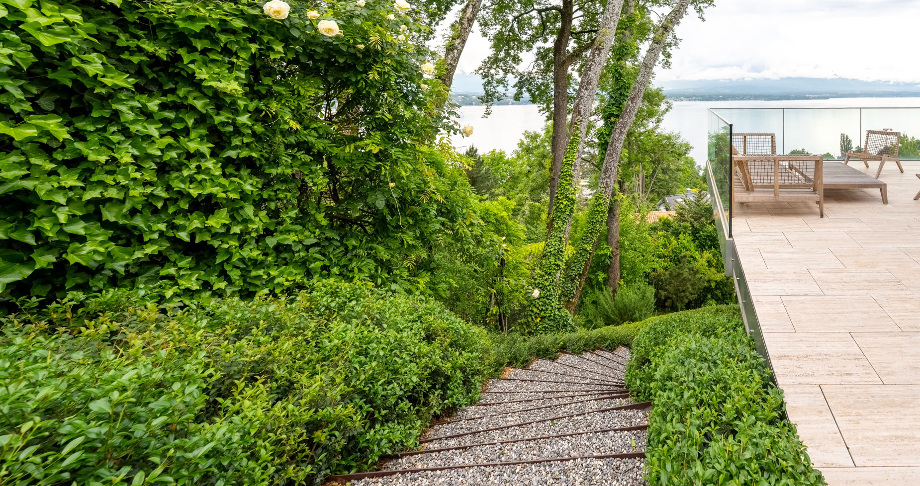 Aménagement jardin Cologny. Escalier en gravier entouré de buissons verts menant à une terrasse avec vue sur un plan d'eau et des arbres.
