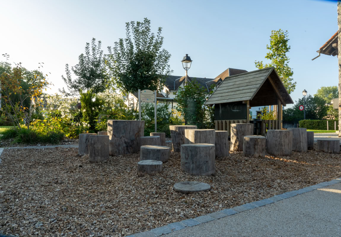 Aire de jeux extérieure avec des structures en bois sur un sol recouvert de copeaux de bois, entourée de verdure et d'arbres, avec un bâtiment en arrière-plan.