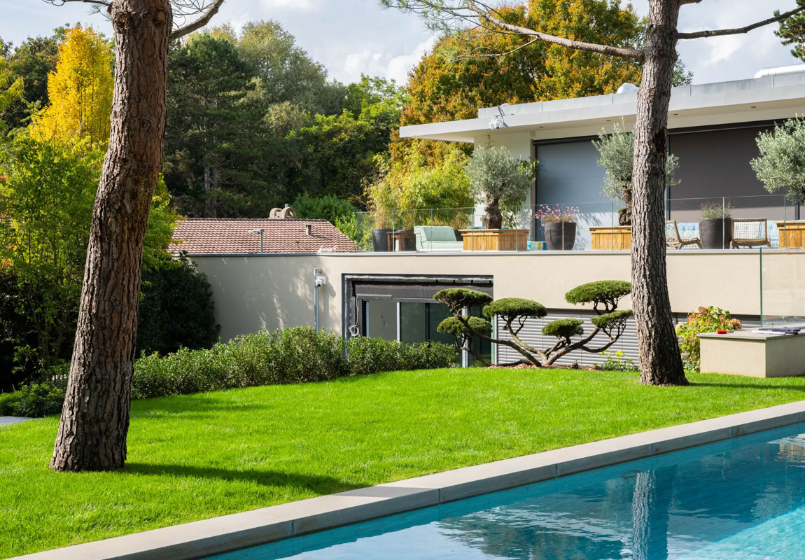 Maison moderne avec terrasse, jardin bien entretenu, piscine au premier plan, arbres et végétation environnante.