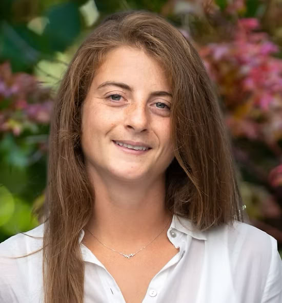 Serena Bondi souriant portant une chemise blanche, avec un fond flou de feuillage coloré.