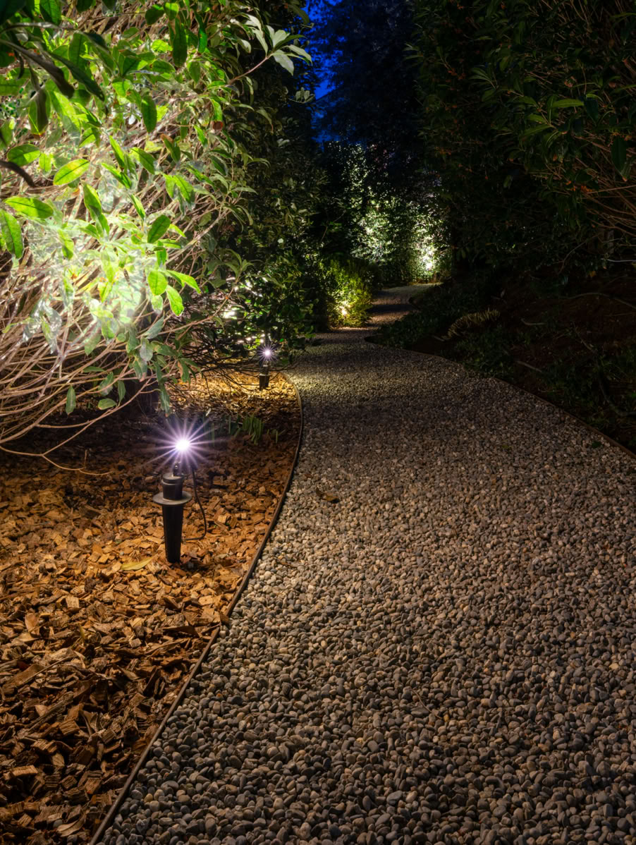Sentier de gravier éclairé par des lampes solaires, entouré de buissons et d'arbres dans un jardin la nuit à Versoix.