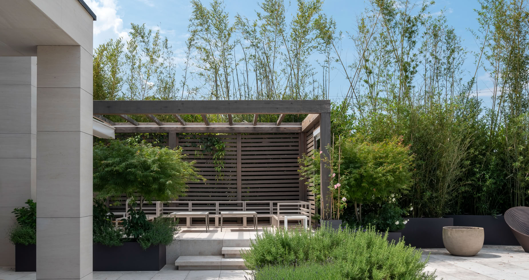 Terrasse extérieure d'un aménagement jardin à Cologny avec pergola en bois, entourée de plantes vertes et de bambous, ciel bleu en arrière-plan.