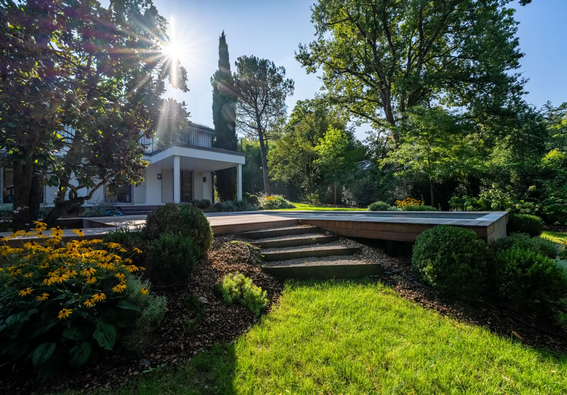 Maison blanche entourée de verdure avec des arbres et des buissons, escalier menant à une terrasse, soleil brillant à travers les arbres.