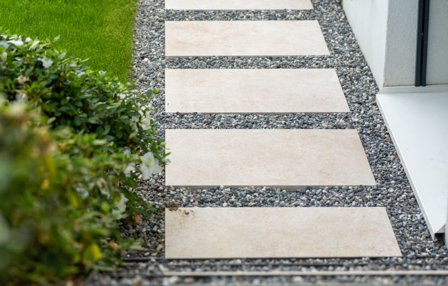 Allée en dalles de pierre entourée de gravier noir, bordée de pelouse verte et de buissons, menant à une porte.
