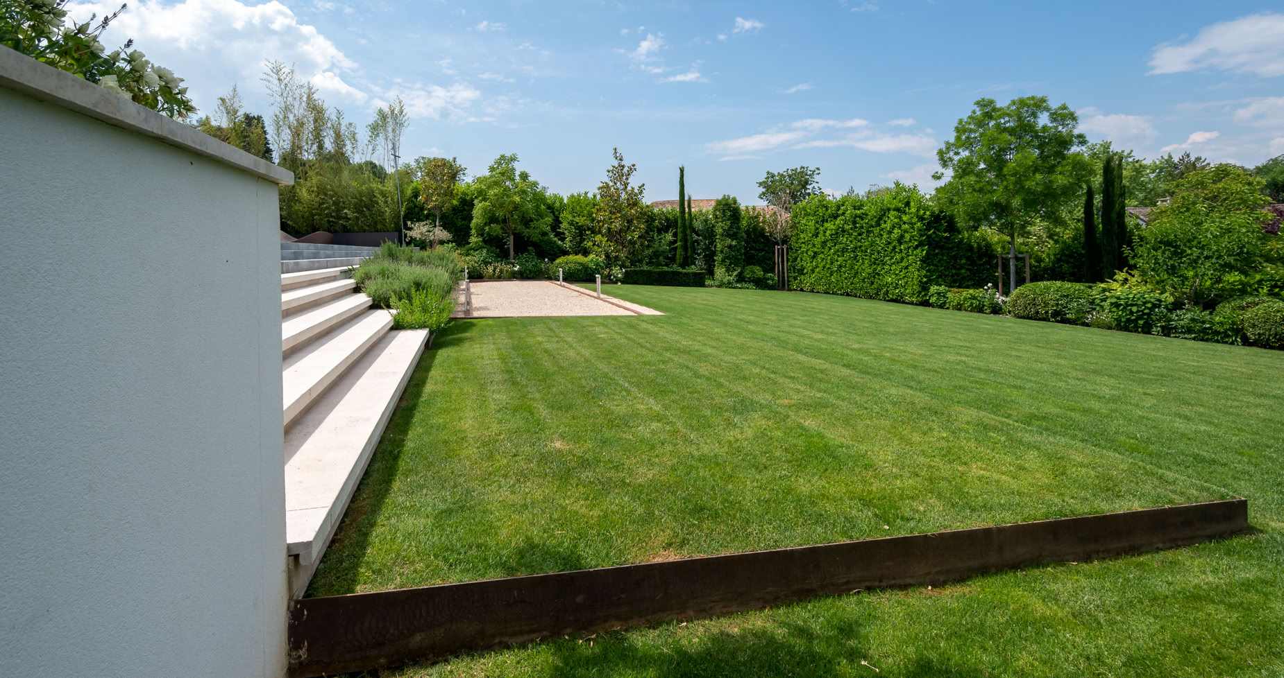 Pelouse verte avec des escaliers blancs à gauche, un terrain de pétanque au centre, arbres et ciel bleu en arrière-plan à Cologny.
