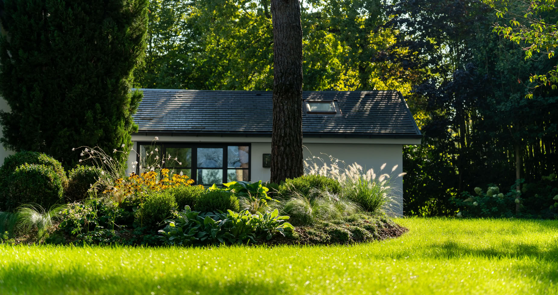 Maison blanche entourée de verdure avec un grand arbre et un jardin fleuri au premier plan.