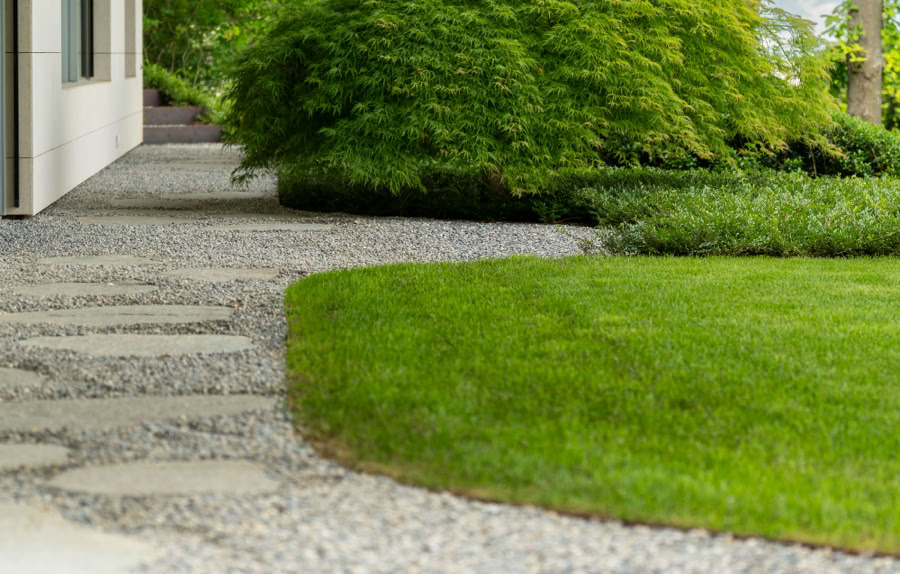 Allée en gravier menant à une maison moderne avec un jardin bien entretenu et des arbustes verts.