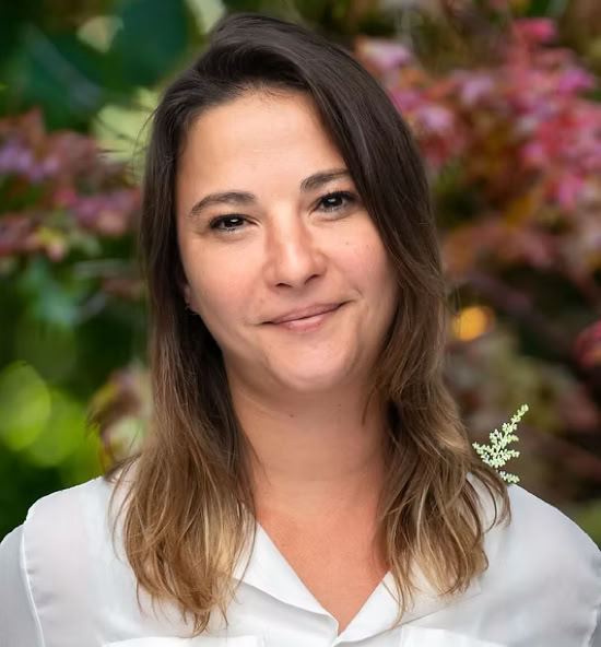 Caroline Hericher souriant portant une chemise blanche, avec un fond flou de feuillage coloré.