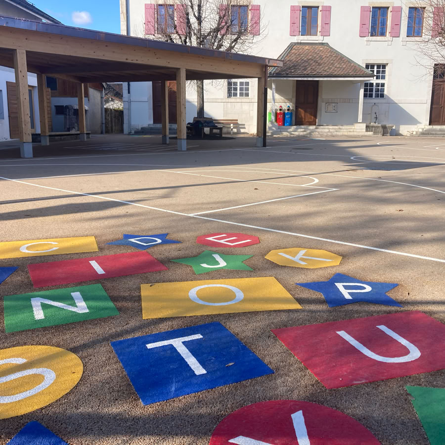 Cour d'école avec des lettres colorées peintes sur le sol, bâtiments en arrière-plan et arbres sans feuilles.