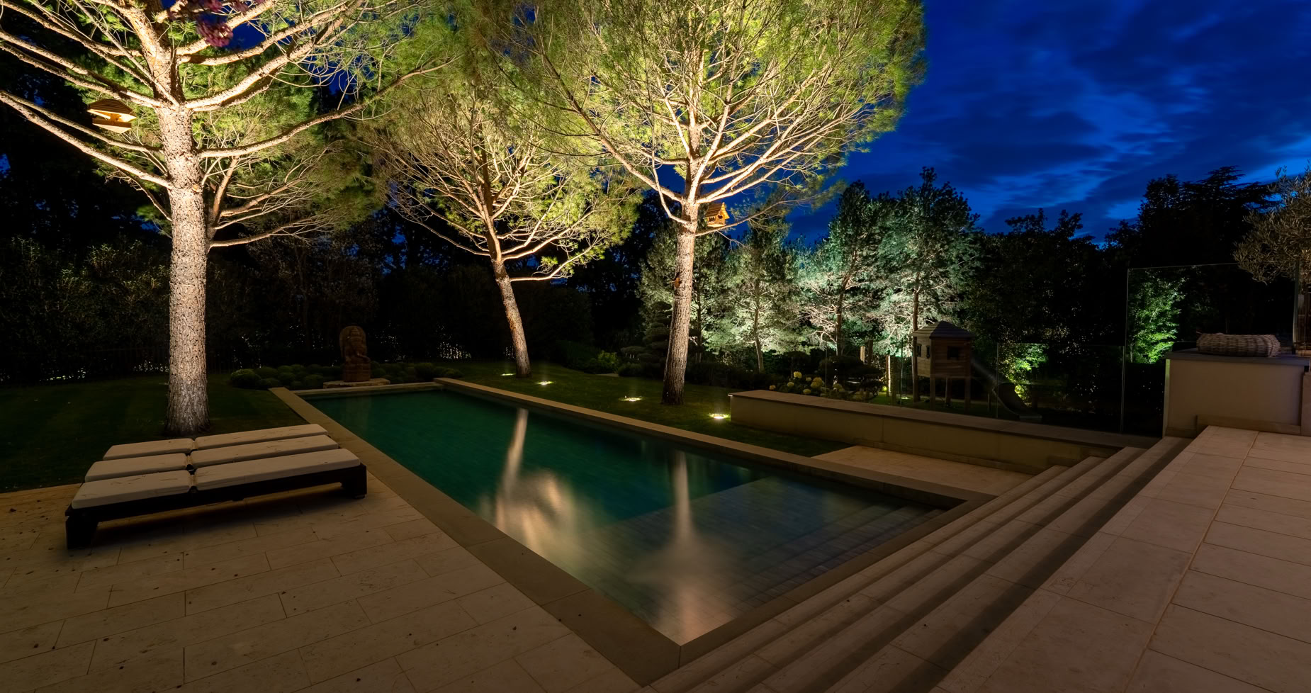 Aménagement jardin à Versoix. Piscine extérieure éclairée la nuit, entourée d'arbres et de végétation, avec des marches en pierre au premier plan.