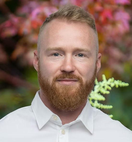 Valentin Perroud souriant portant une chemise blanche, avec un fond flou de feuillage coloré.