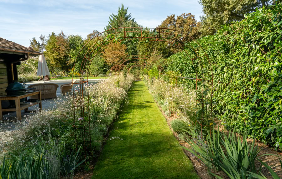 Allée de jardin bordée de plantes et d'arbustes, avec une pelouse verte au centre, entourée de végétation dense et d'un cabanon de jardin à gauche à Collonge-Bellerive.