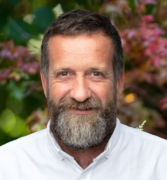 Matthias Deruette souriant portant une chemise blanche, avec un fond flou de feuillage coloré.