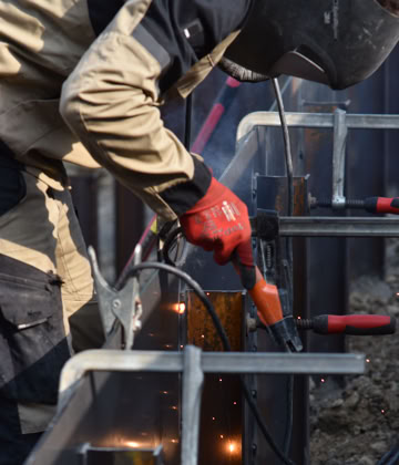 Paysagiste Pottu & Seitz portant un casque de protection et des gants rouges, soudant des pièces métalliques.